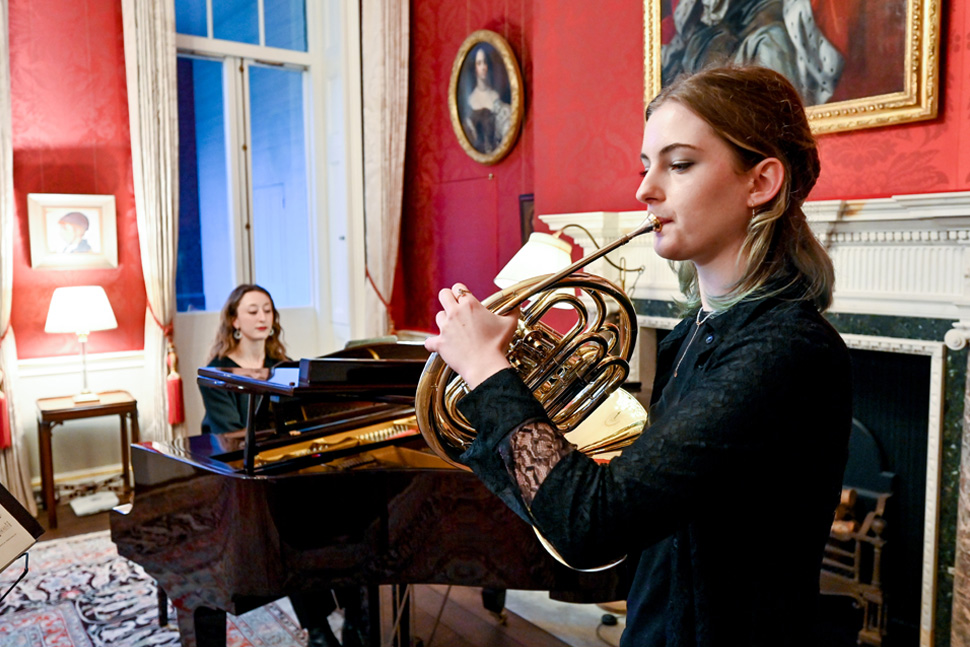 Students wearing smart attire, performing a duet (french horn and piano) in a red room decorated with artwork.
